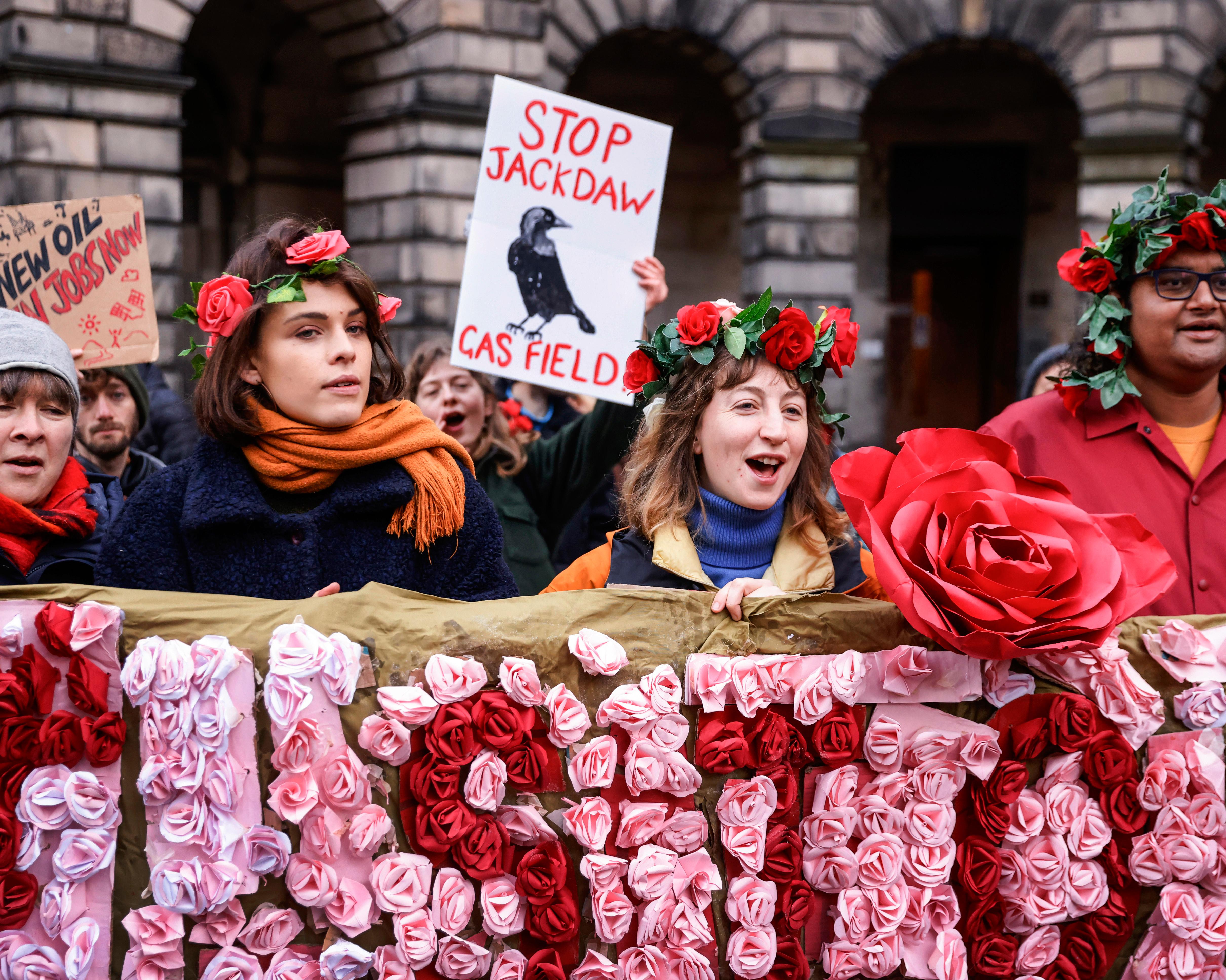 Protesters wearing rose garlands hold a “Stop Jackdaw Gas Field” sign outside a stone building.