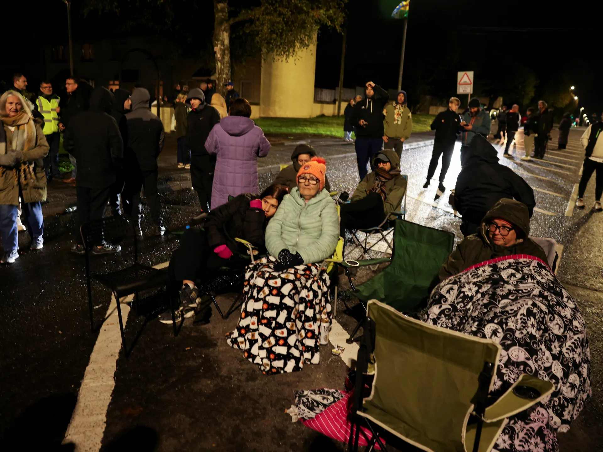 People sit in camping chairs and gather on a wet road at night, wrapped in blankets and coats.