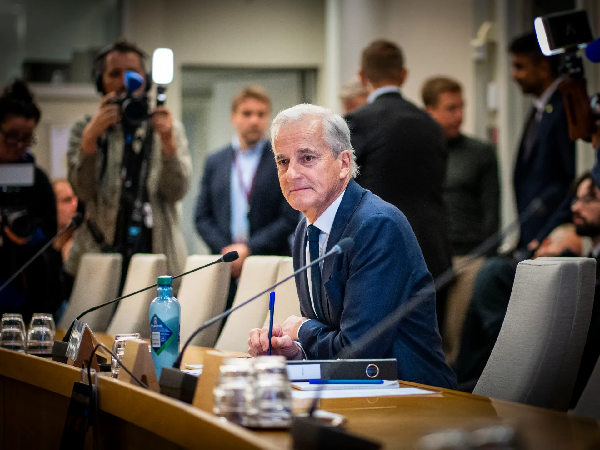White-haired man in a navy suit sits at a conference table holding a pen as journalists film nearby.