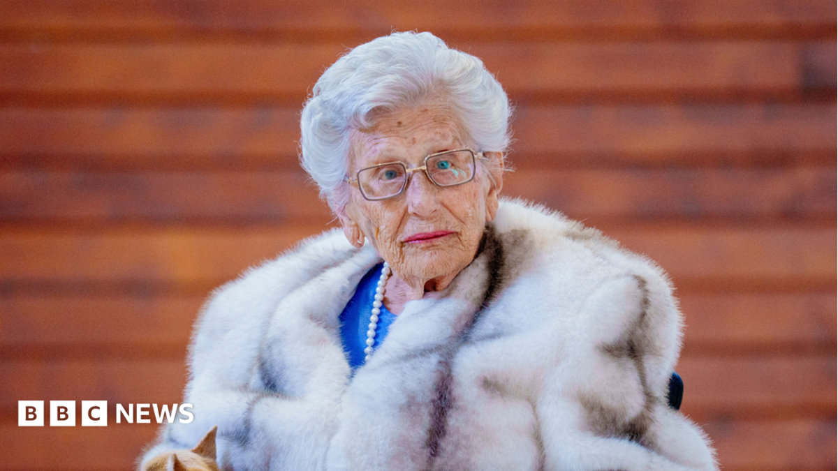 Elderly woman wearing glasses, pearls and a white fur coat seated against a wooden wall