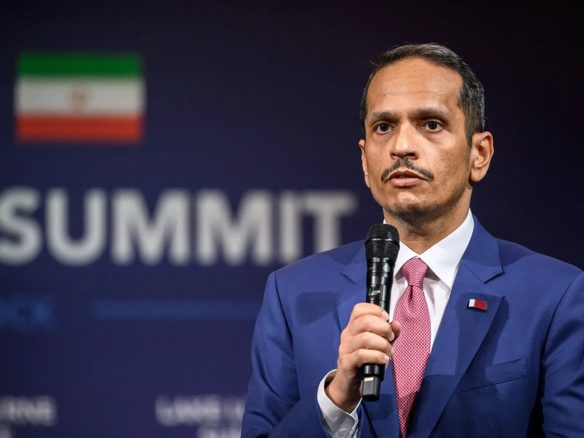 A man in a blue suit speaks into a microphone onstage, with an Iranian flag behind him.