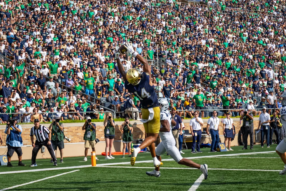 Notre Dame football player leaps to catch a pass near the end zone as fans watch.