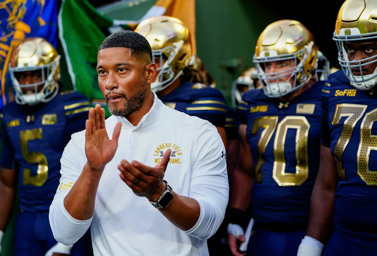 Football coach claps in front of helmeted Notre Dame players in a locker room