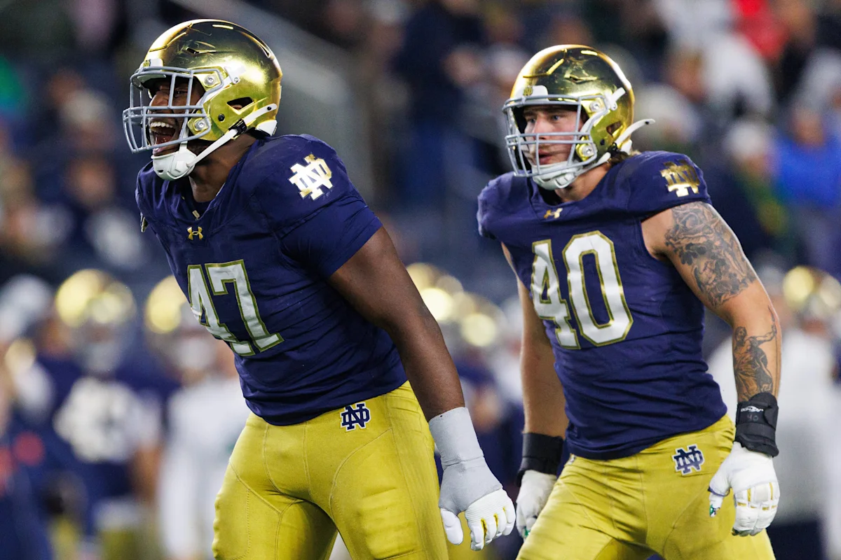Two Notre Dame football players in helmets and uniforms walk across the field during a game.