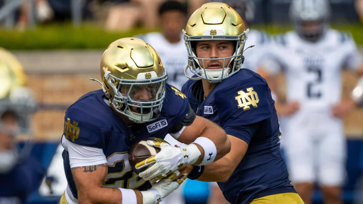 Notre Dame football players in gold helmets and navy uniforms execute a handoff during a game.