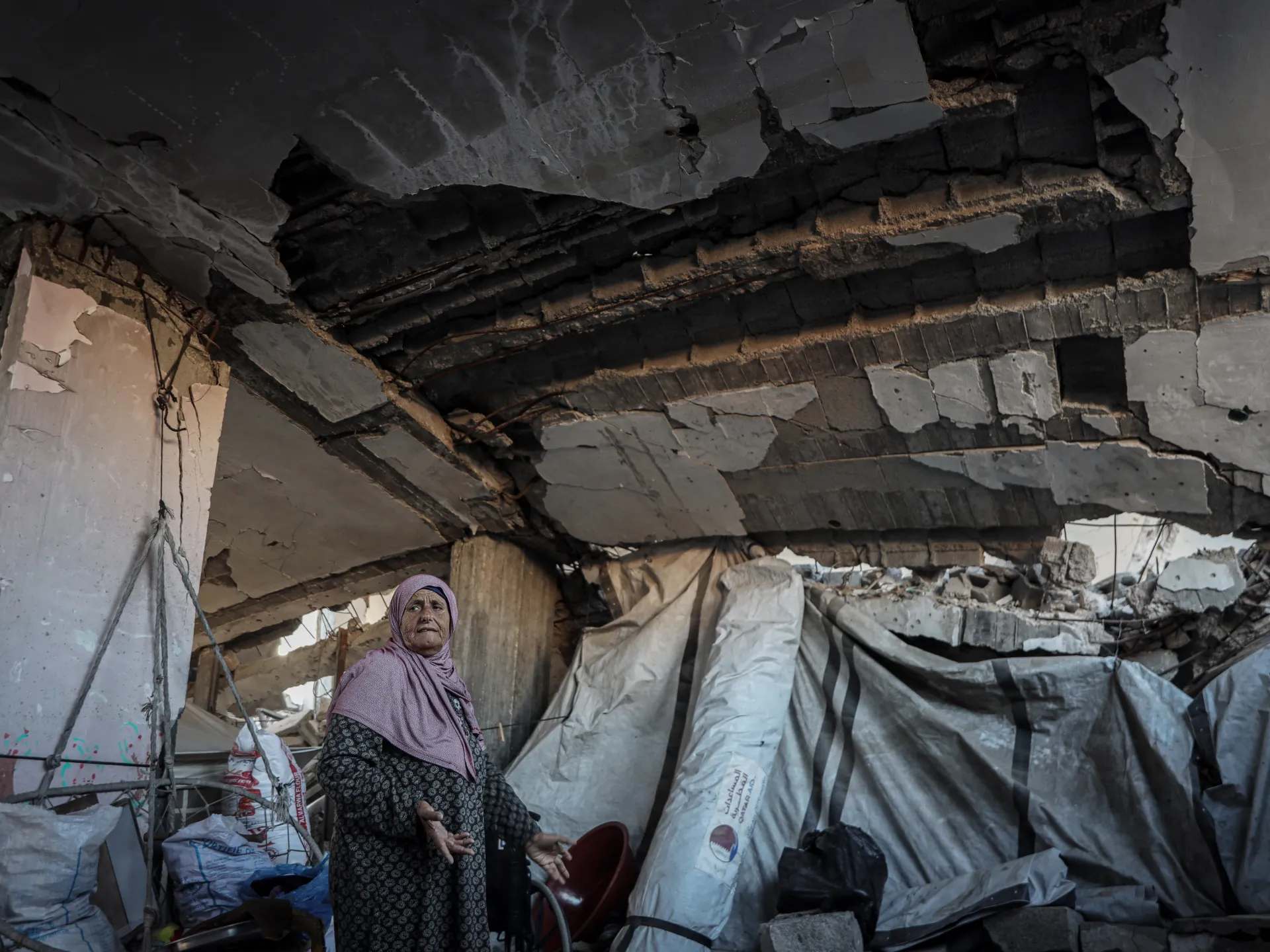 An older woman stands amid sacks and debris beneath a severely cracked, partially collapsed concrete ceiling.