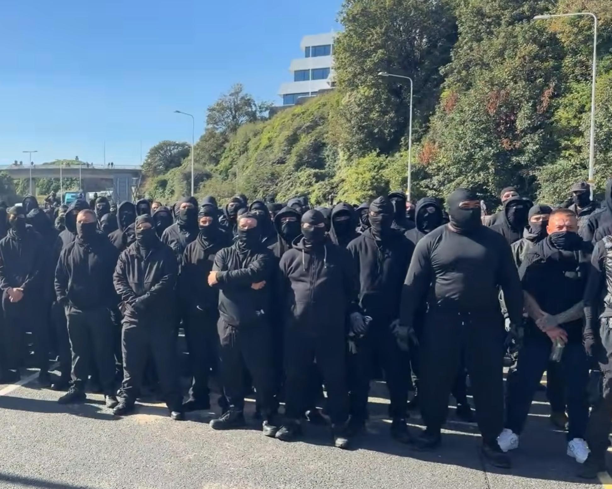 Large group of people dressed in black, many with face coverings, standing on a city road.