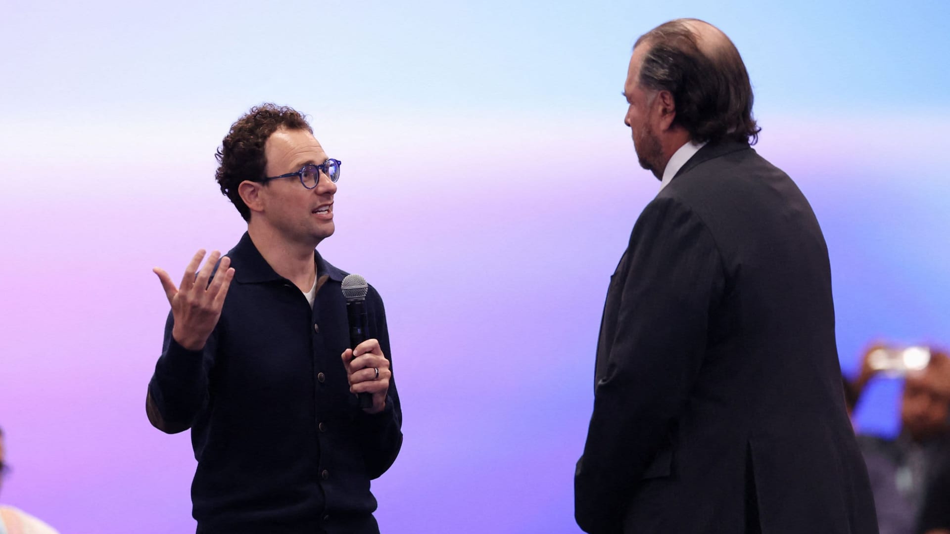Two men speak onstage, one holding a microphone and gesturing, against a purple-blue backdrop.
