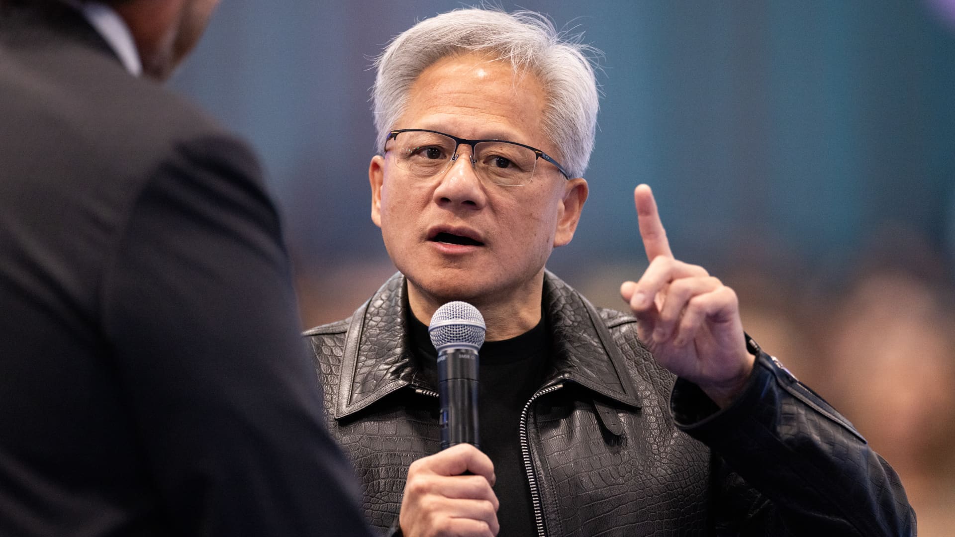 Silver-haired man in glasses speaks into a microphone while raising one finger.