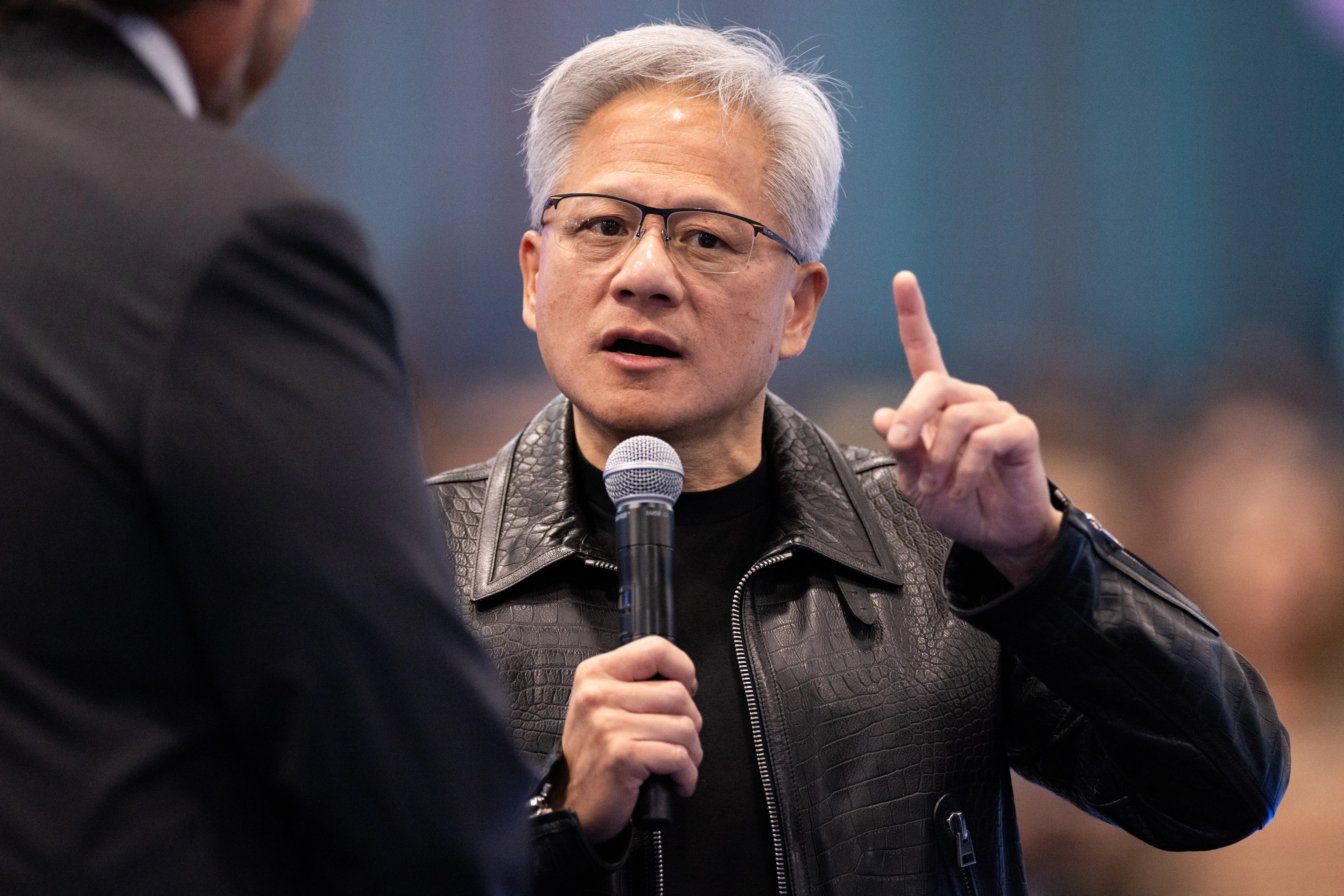 Gray-haired man in glasses speaks into a microphone and raises one finger, wearing a black leather jacket.