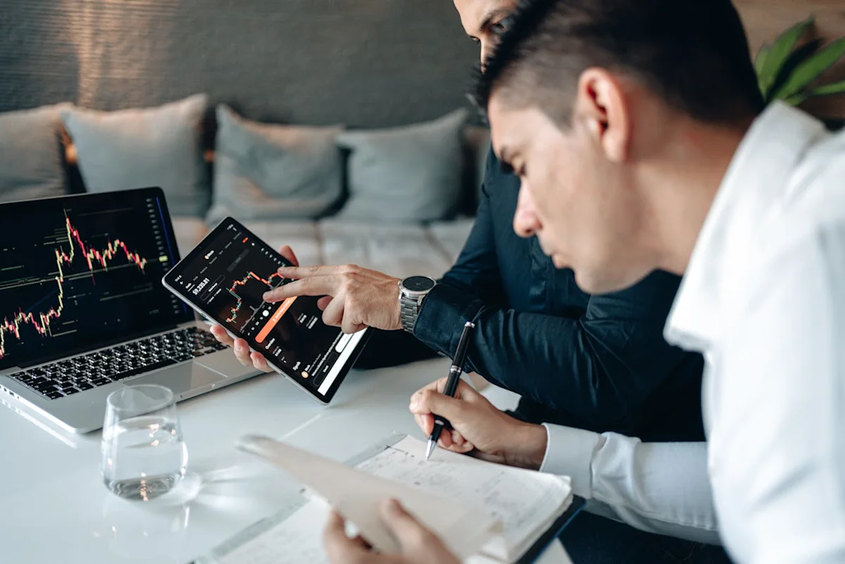 Two people review financial charts on a laptop and tablet at a table.