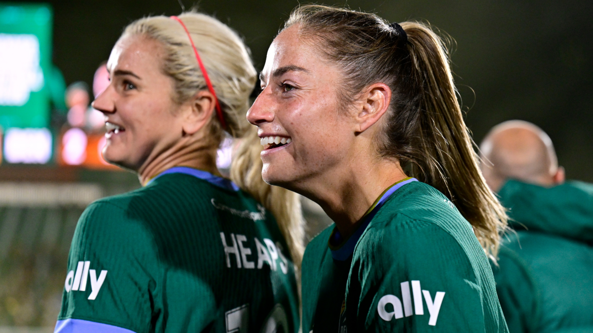 Two women in green soccer jerseys smile during a match, with another person visible in the background.