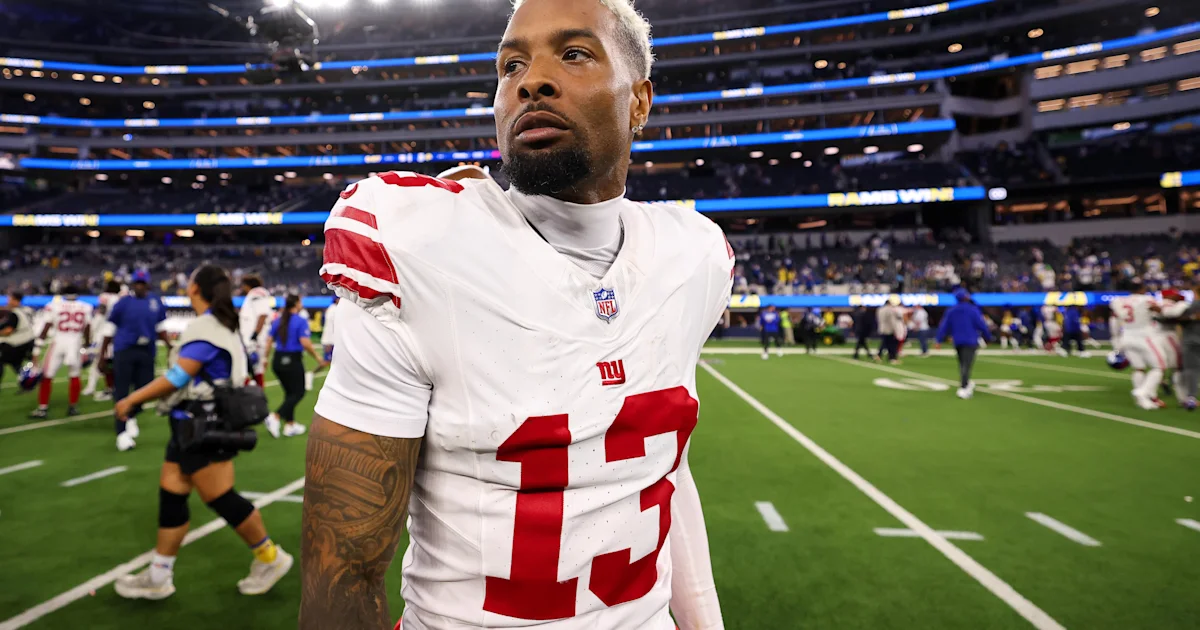 A Giants player wearing number 13 stands on the field in a white uniform as others warm up.