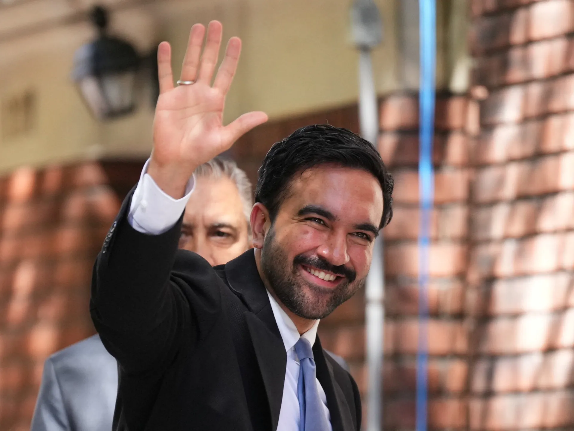 Smiling man in a dark suit waves toward the camera with a blurred crowd behind him