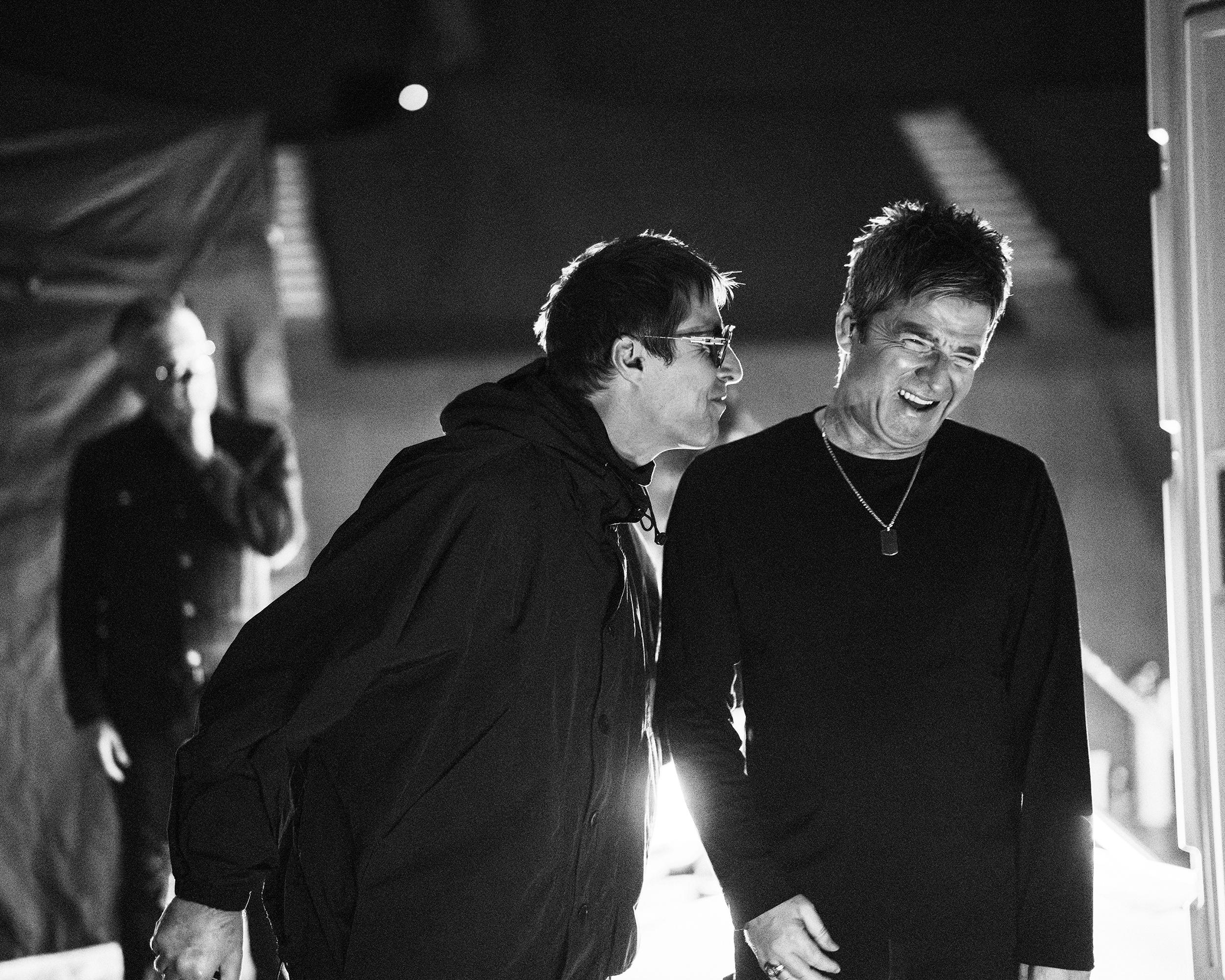 Two men laugh together backstage, with a blurred figure and stage lighting visible behind them.