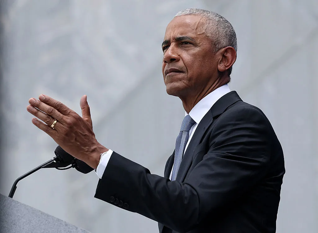 Barack Obama gestures while speaking at a podium, wearing a dark suit and blue tie.
