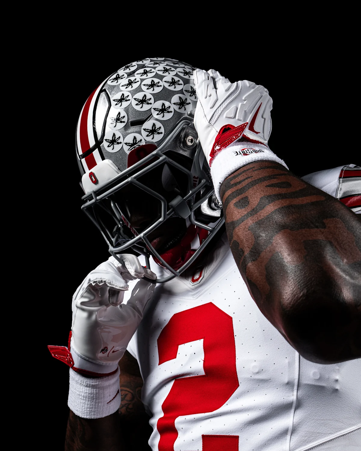 Ohio State football player in white uniform and helmet adjusts facemask against black background
