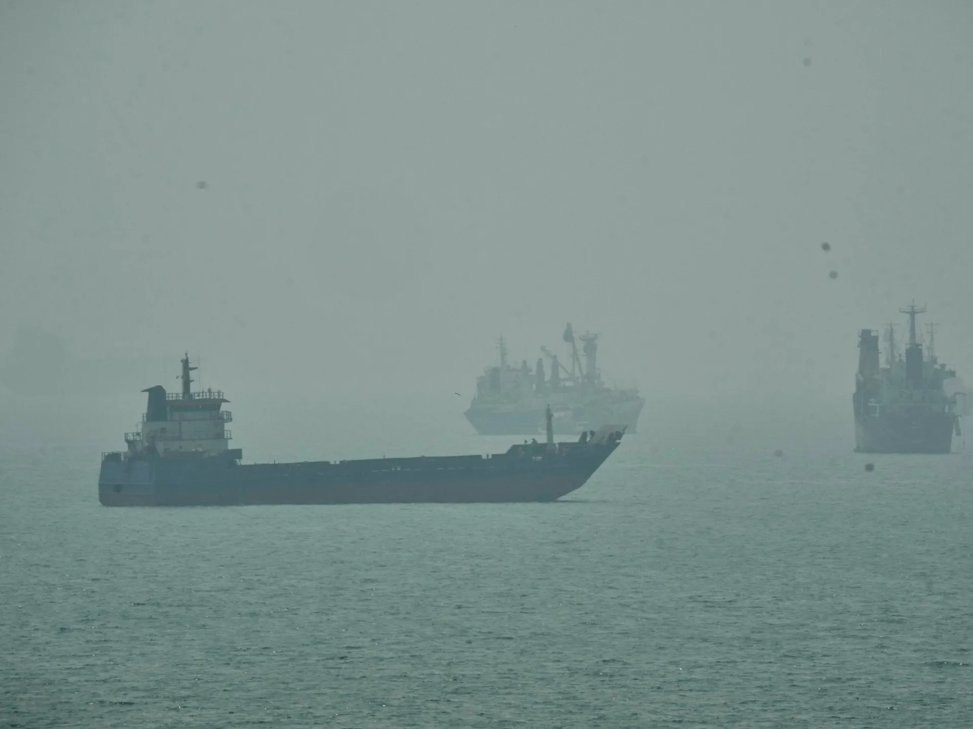 Several ships sit on hazy water, with a long dark vessel in the foreground.