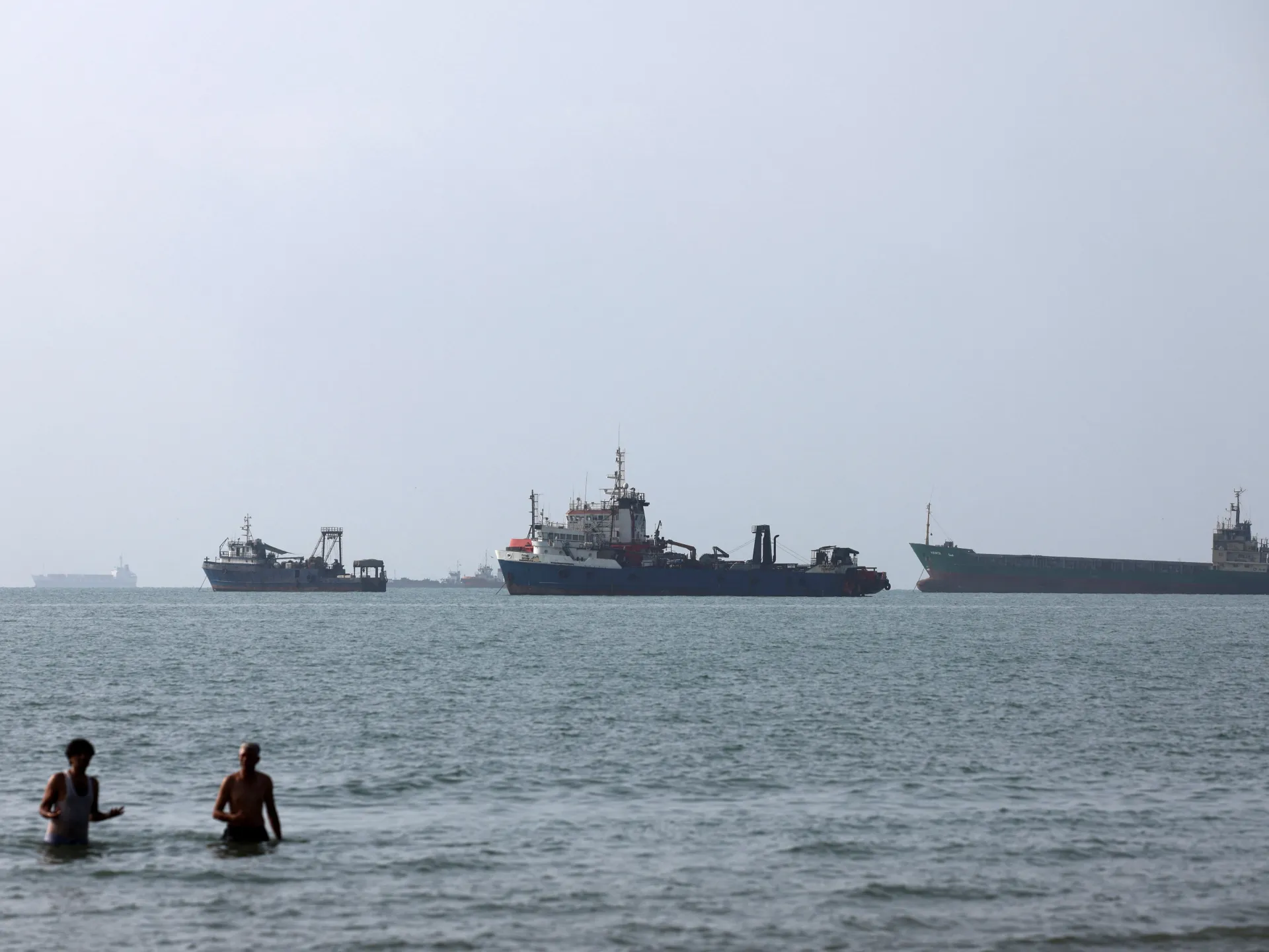 Several ships sit offshore while two people wade in calm seawater under a hazy sky.