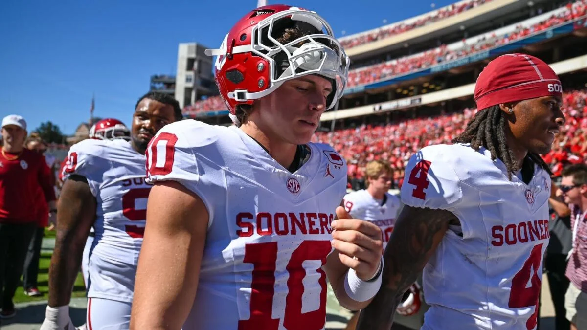 Oklahoma players in white uniforms walk off the field beneath a sunny stadium crowd.