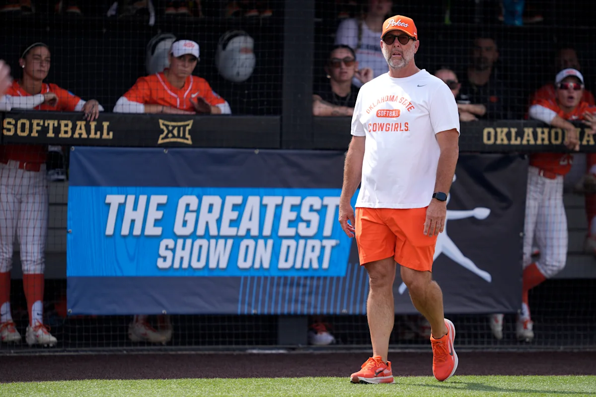 Oklahoma State softball coach walks across the field wearing orange shorts and a white team shirt.