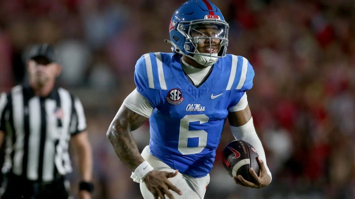 Ole Miss player in a blue number 6 uniform holds a helmet beside a blurred referee.