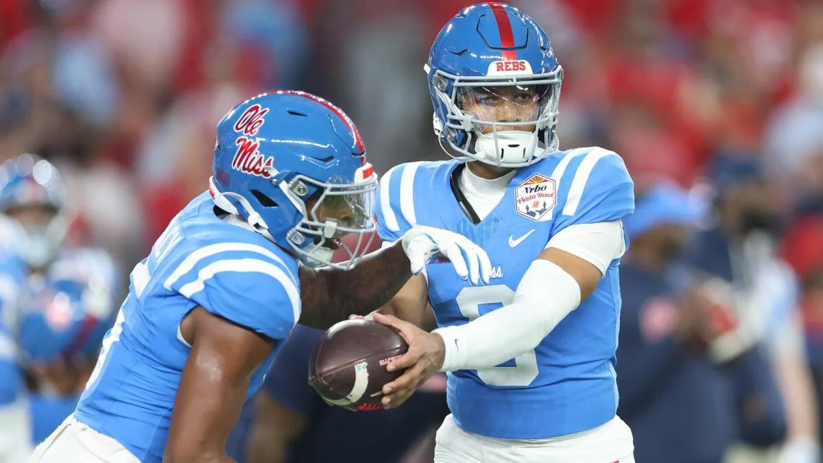 Two Ole Miss football players in blue uniforms execute a handoff during a game.