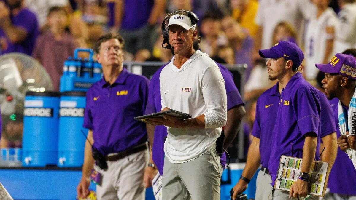 LSU coach in white headset stands on sideline with staff and players during a football game.