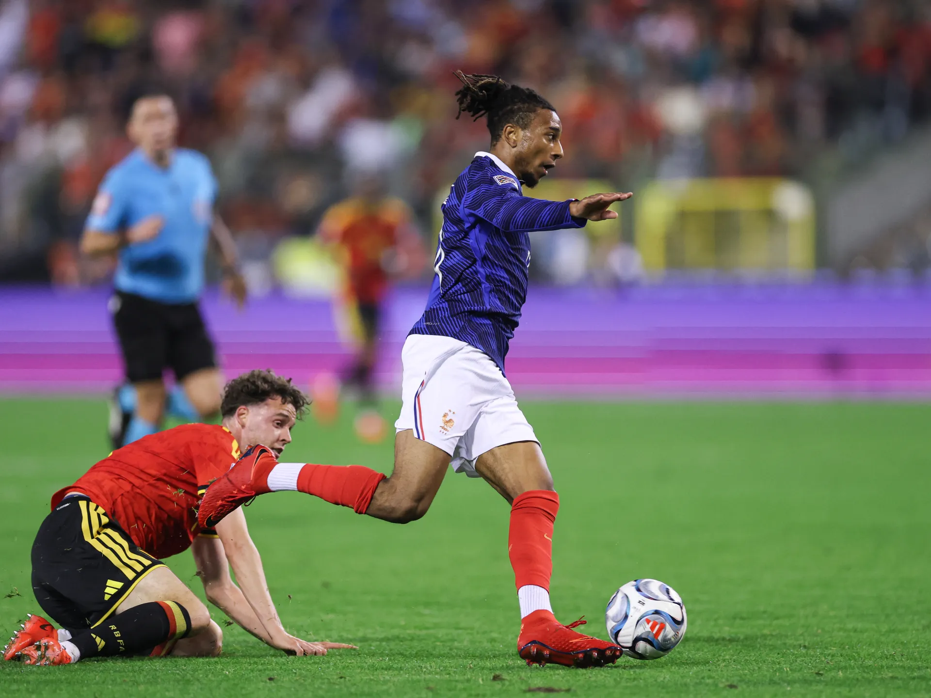 France player in blue runs with the ball as a Belgium player in red falls behind him.