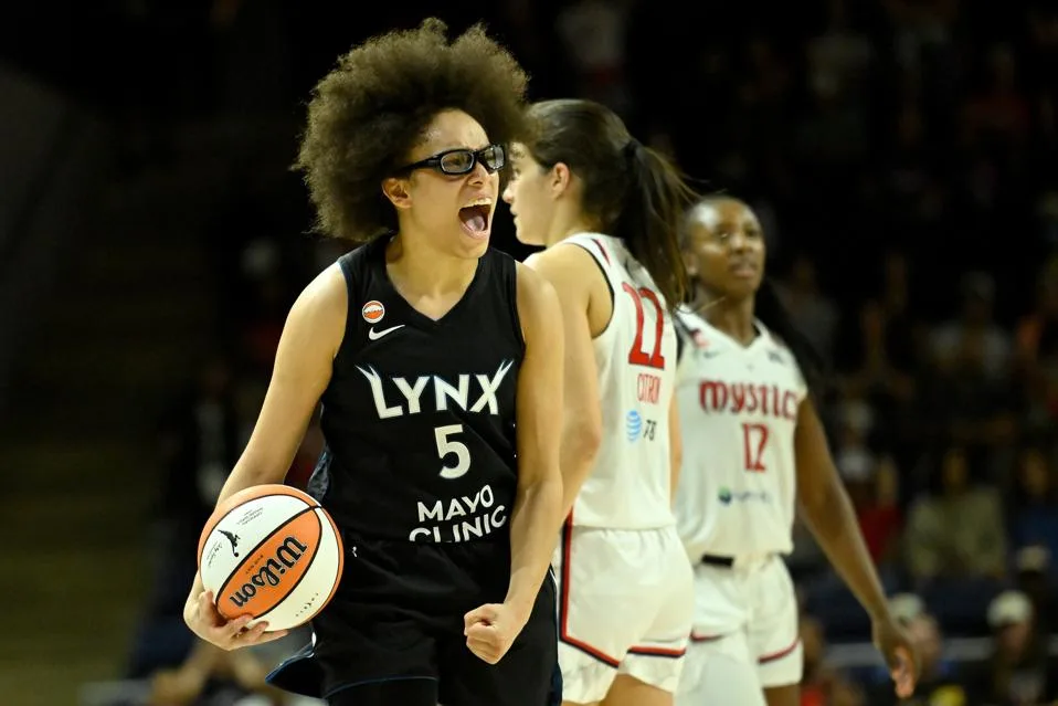Basketball player in a black Lynx jersey shouts while holding a ball near two players in white uniforms.
