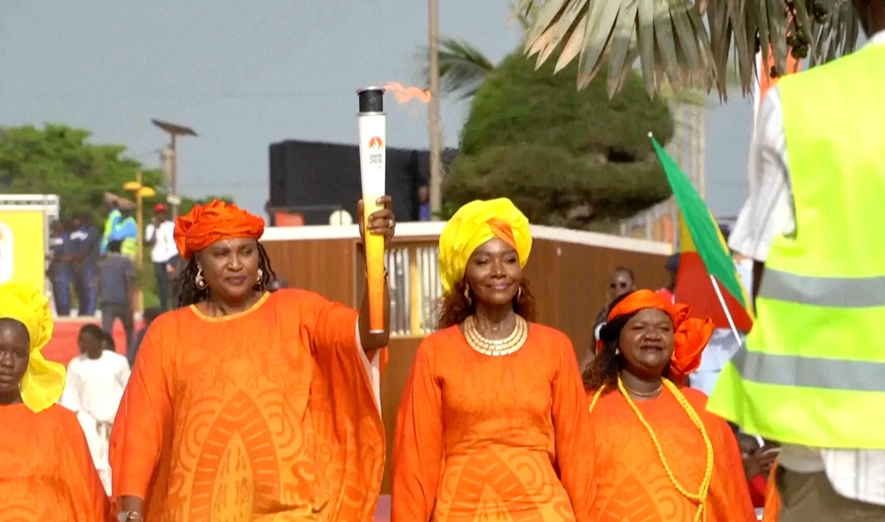 People in orange clothing walk beside a torch during a public outdoor procession.