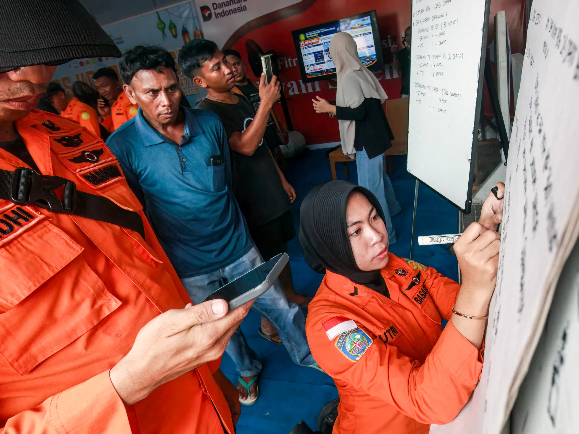 Rescue workers and civilians gather around whiteboards inside a crowded emergency response area.