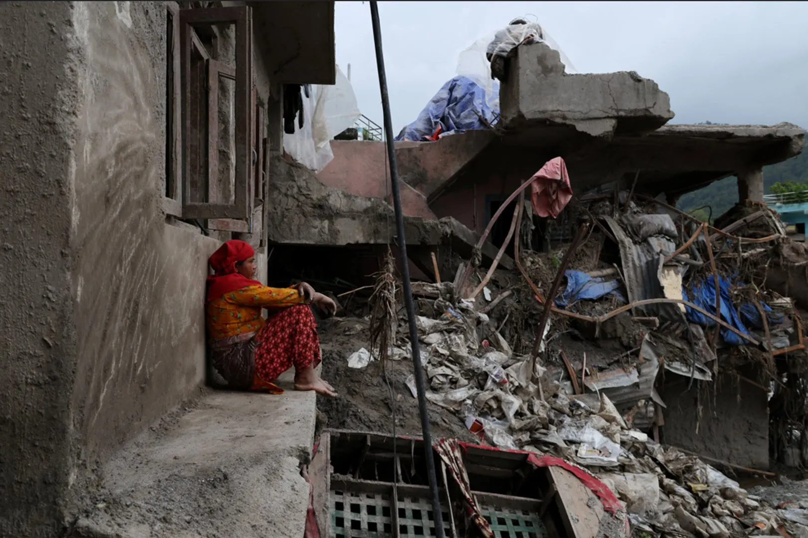 A woman in a red headscarf sits beside a collapsed building surrounded by rubble.
