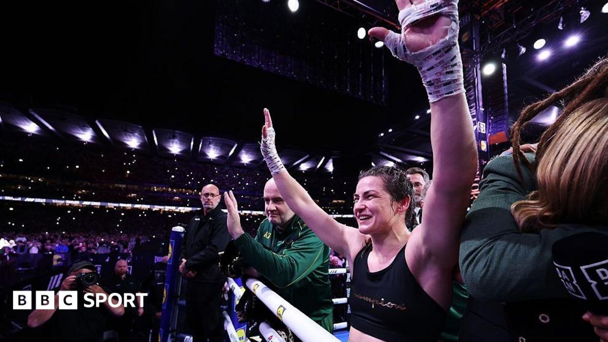 A female boxer raises her taped fists as supporters celebrate ringside in a packed arena.