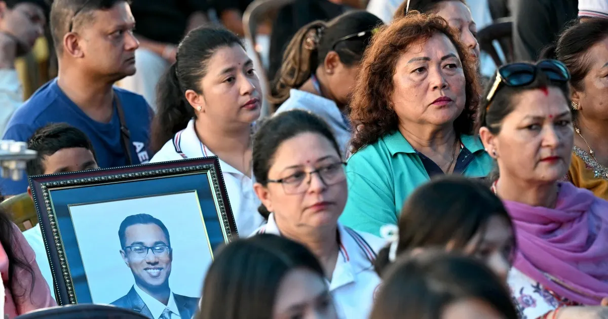 Crowd gathered outdoors, with mourners holding a framed portrait of a smiling man.