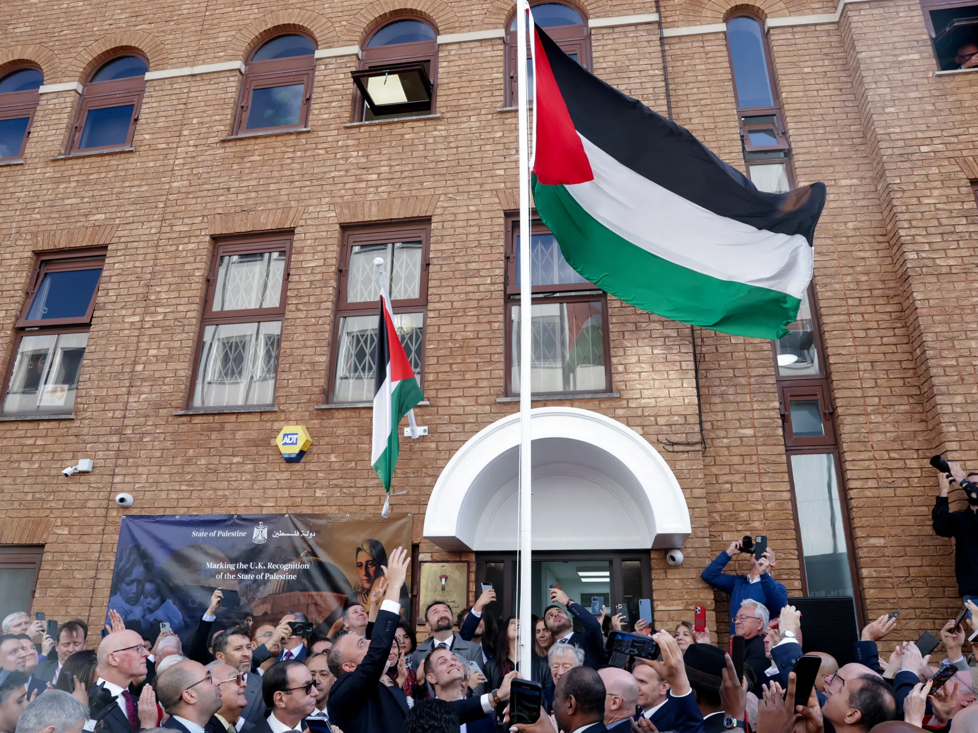 A large Palestinian flag flies above a crowd gathered outside a brick building with a Palestinian statehood banner.