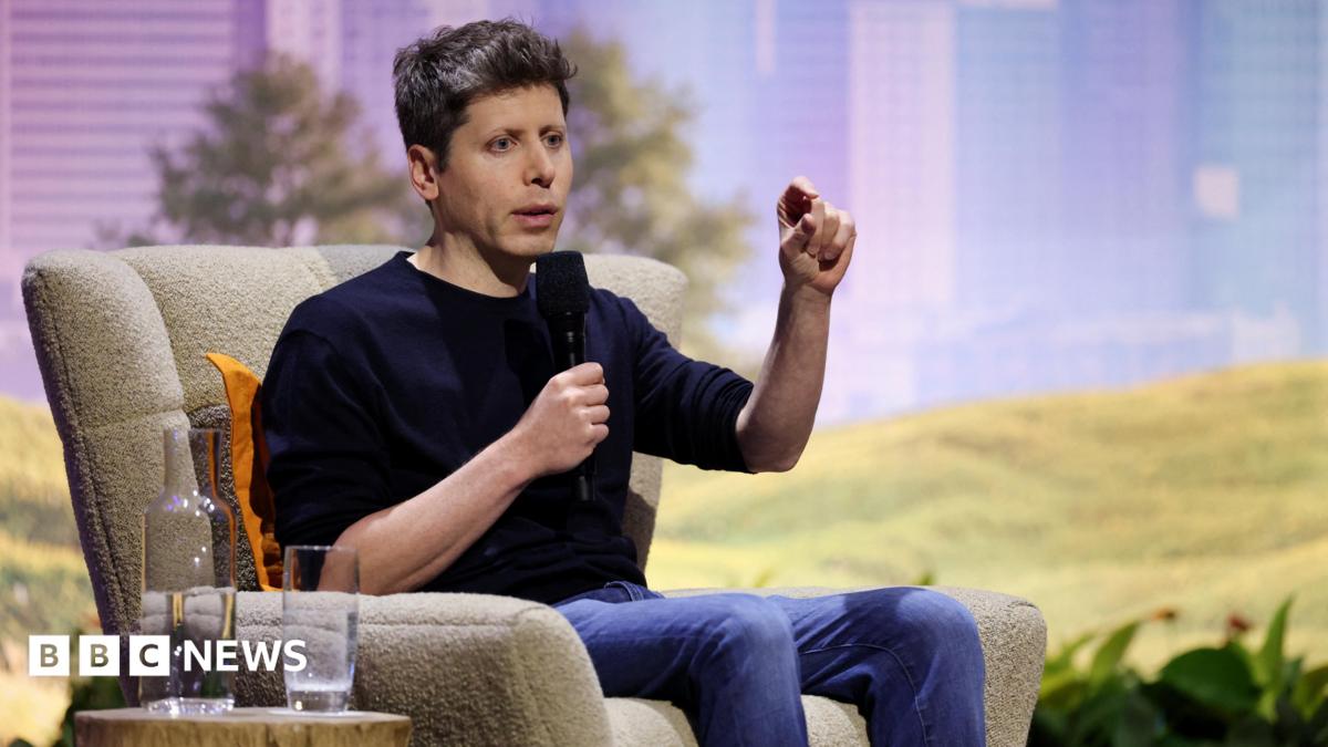 Man speaking into a microphone while seated in an armchair, raising one hand.