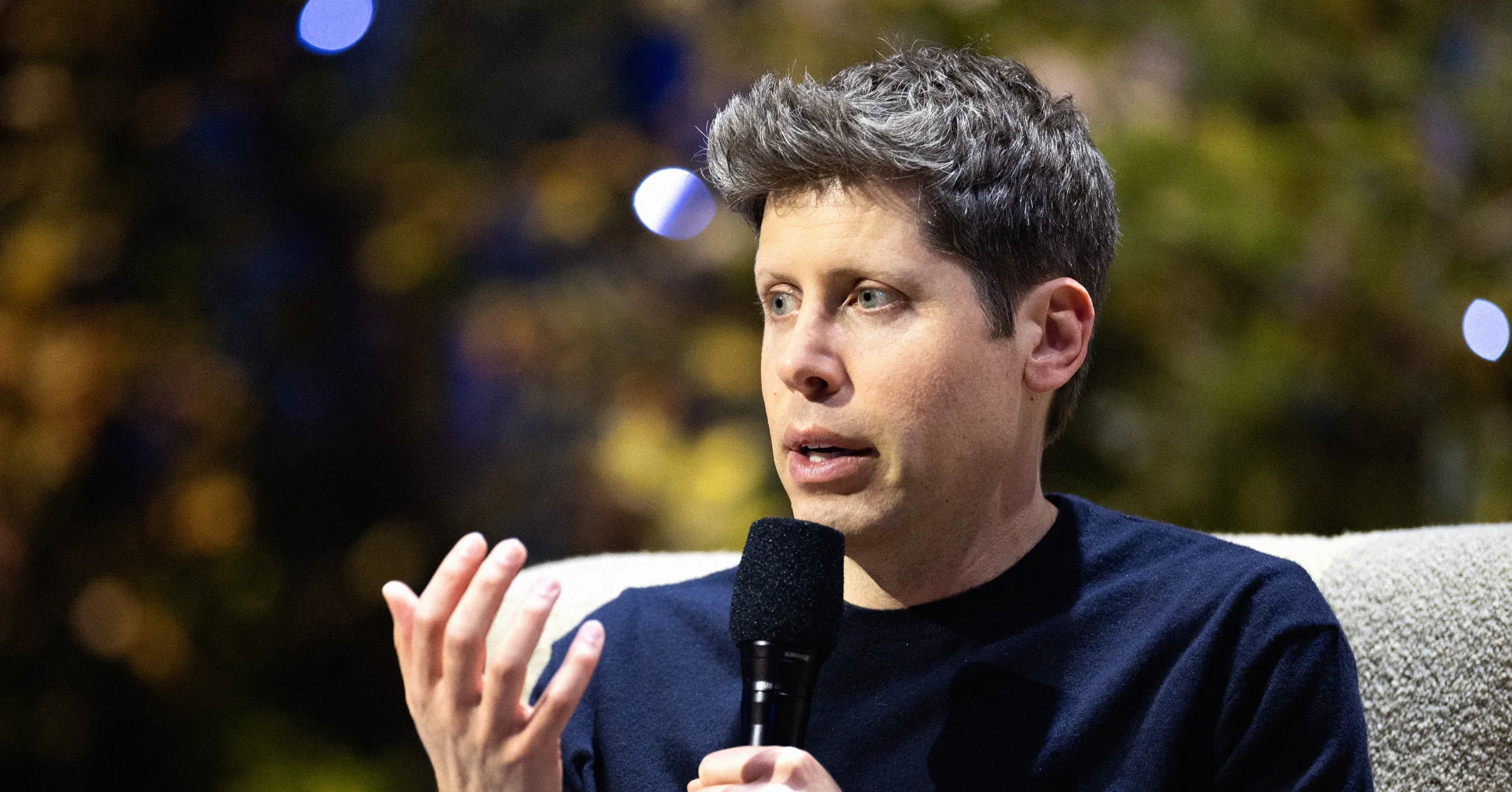 Man holding a microphone and gesturing while speaking in a dimly lit setting