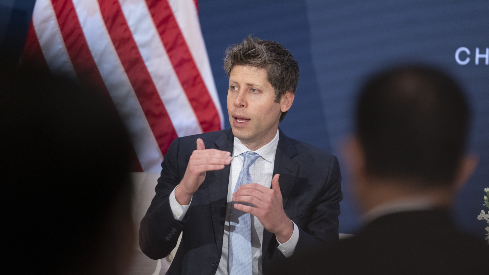 A man in a dark suit gestures while speaking onstage, with an American flag behind him and audience silhouettes in front.