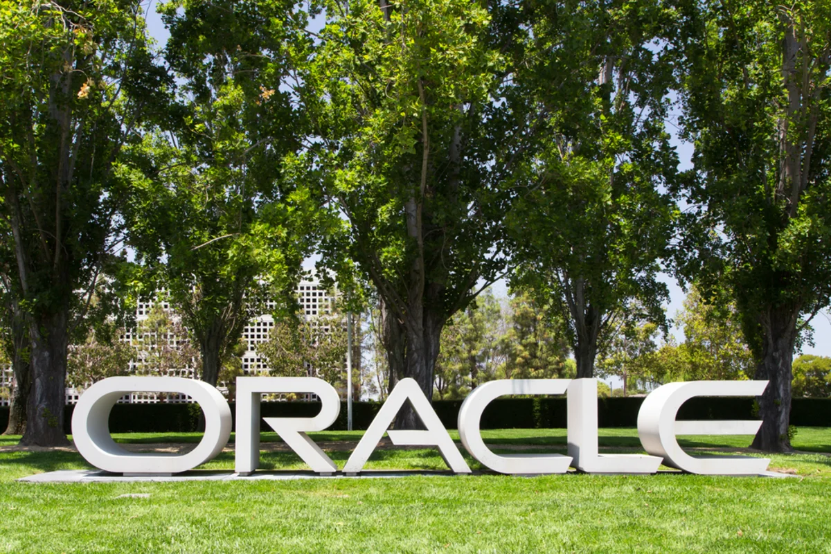 White ORACLE letters displayed on a lawn beneath leafy trees and beside office buildings.