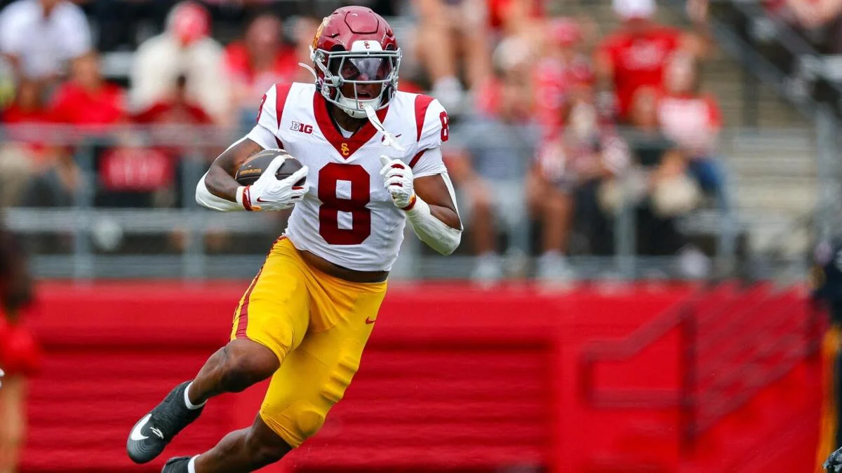 USC player in a white number 8 jersey runs with the football against a blurred stadium crowd.