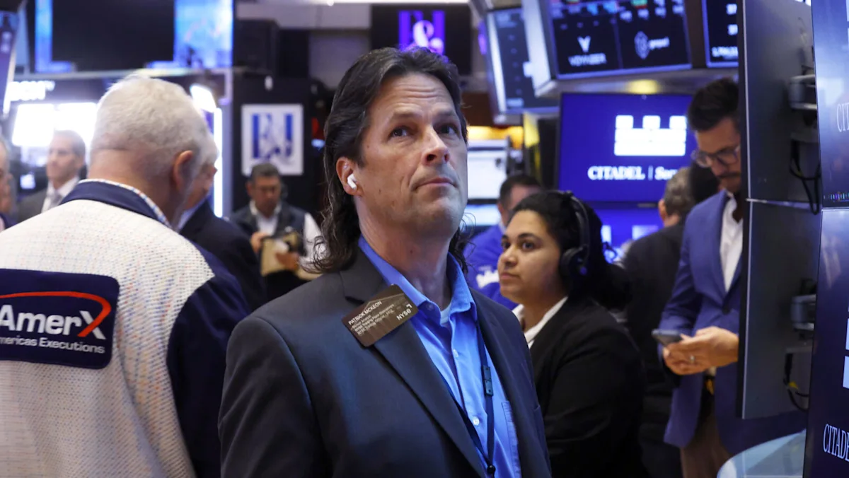 Man in suit wearing an earpiece stands among traders and electronic market screens.