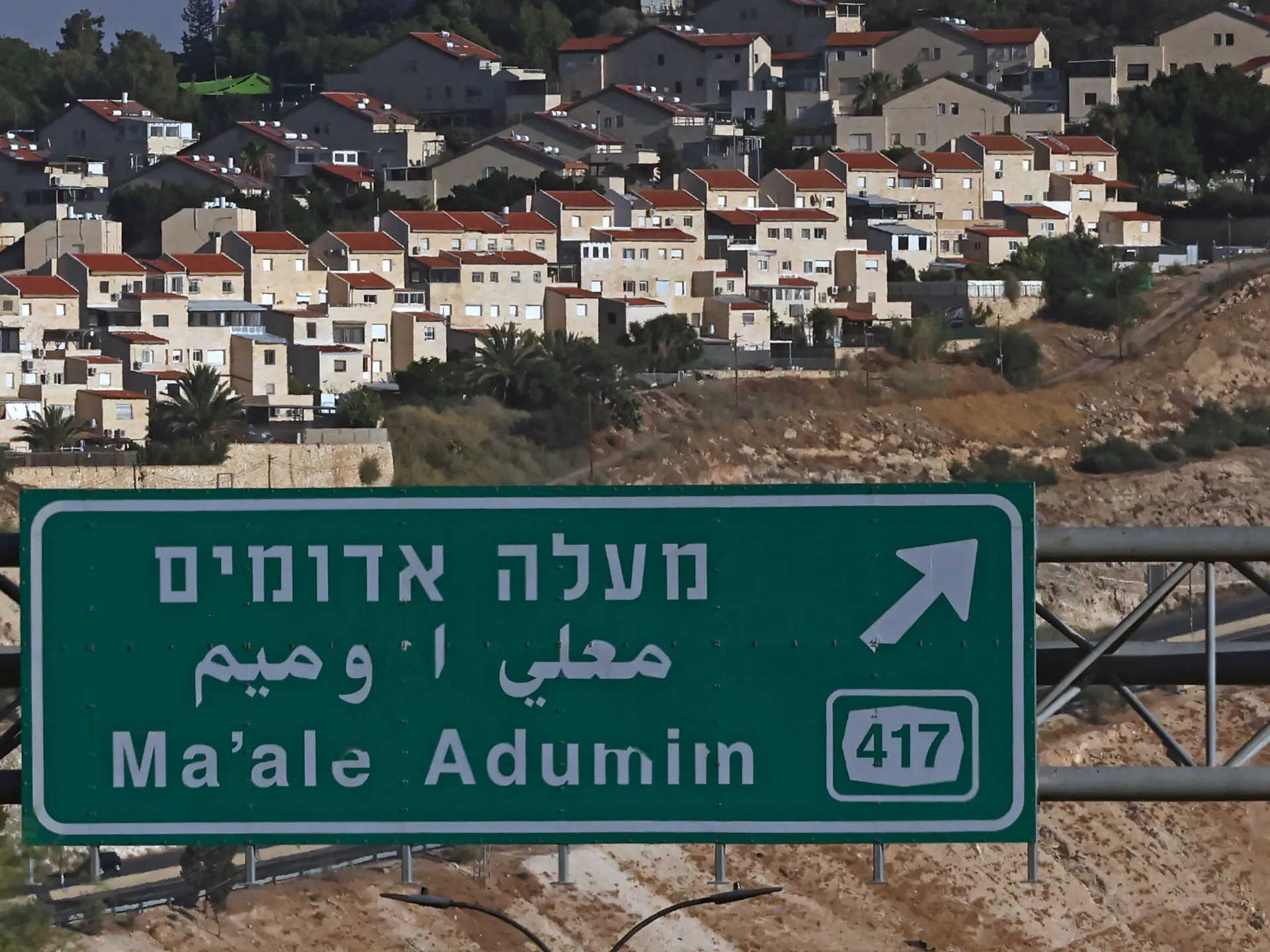 Road sign for Ma'ale Adumim beside densely built hillside homes and arid terrain.
