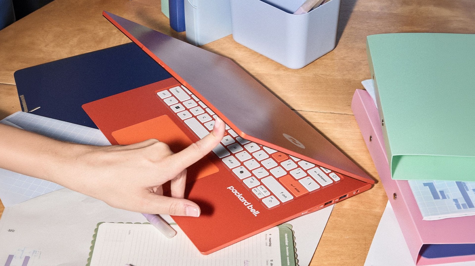 Hand opens a red Packard Bell laptop surrounded by colorful stationery on a wooden desk.