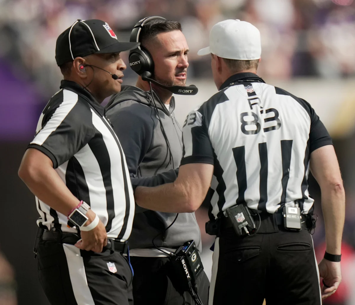 Three football sideline personnel confer during a game, with one wearing a headset and two in striped uniforms.