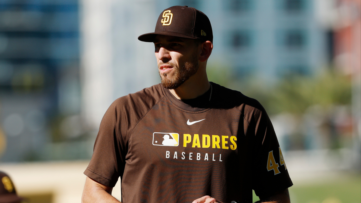 A man in a Padres cap and brown baseball shirt stands outdoors in bright sunlight.
