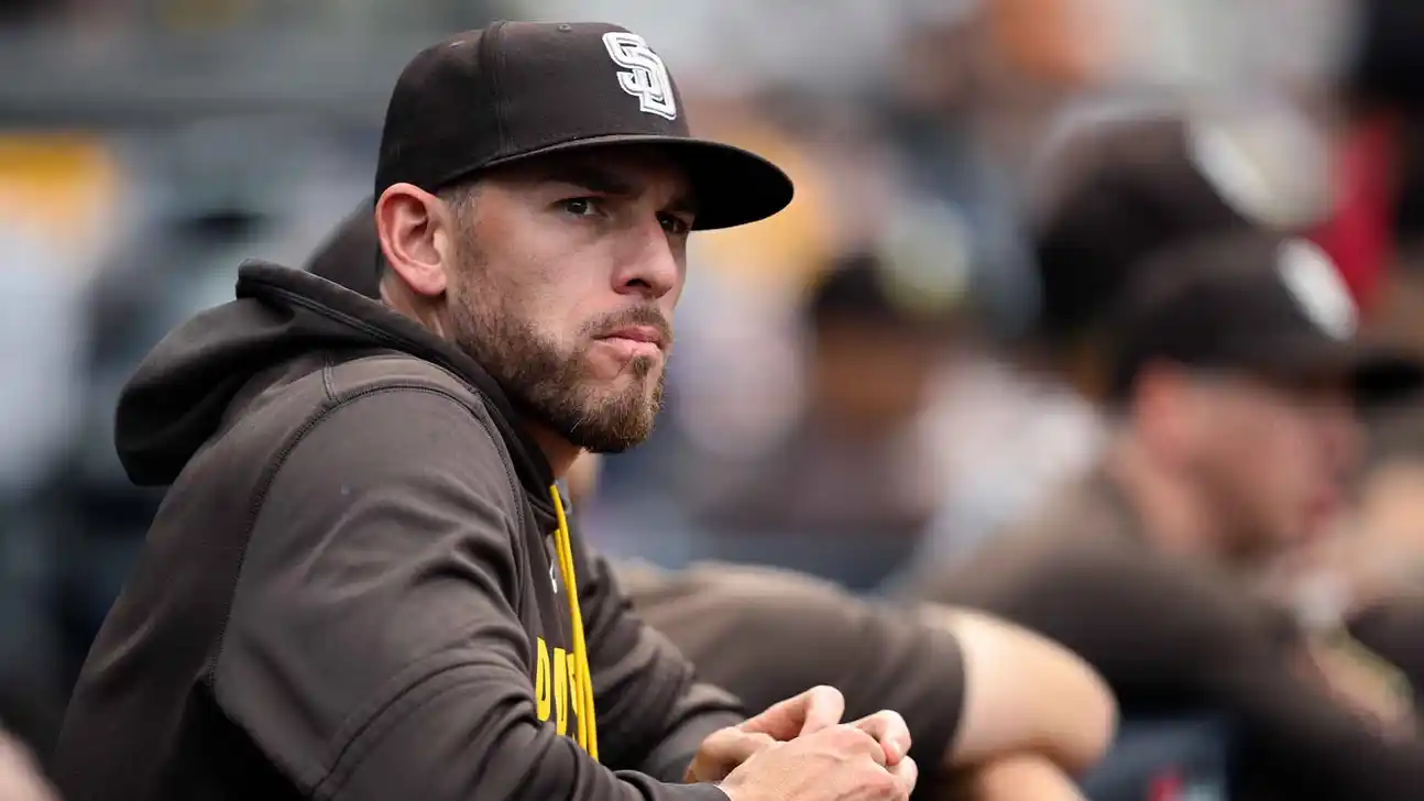 A bearded man in a Padres cap and hoodie sits with his hands clasped near a dugout.