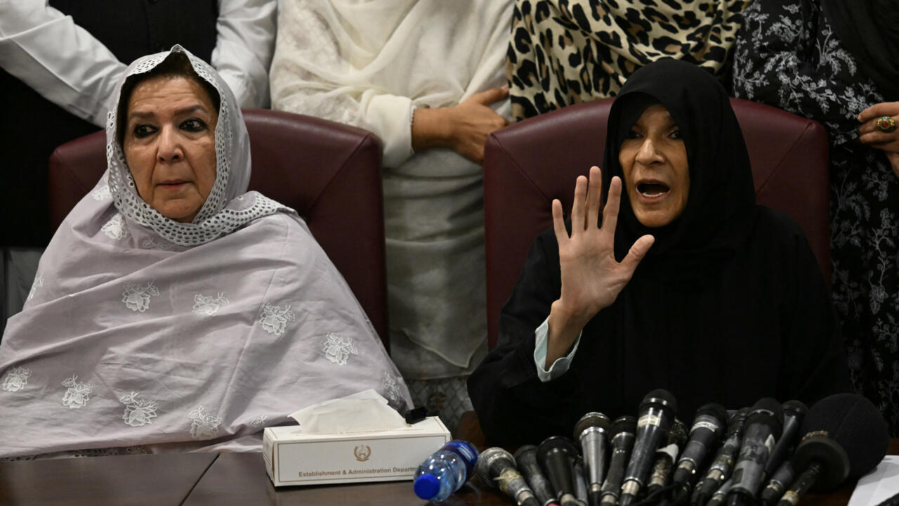 Two women in head coverings sit before microphones, with one raising her hand while speaking.