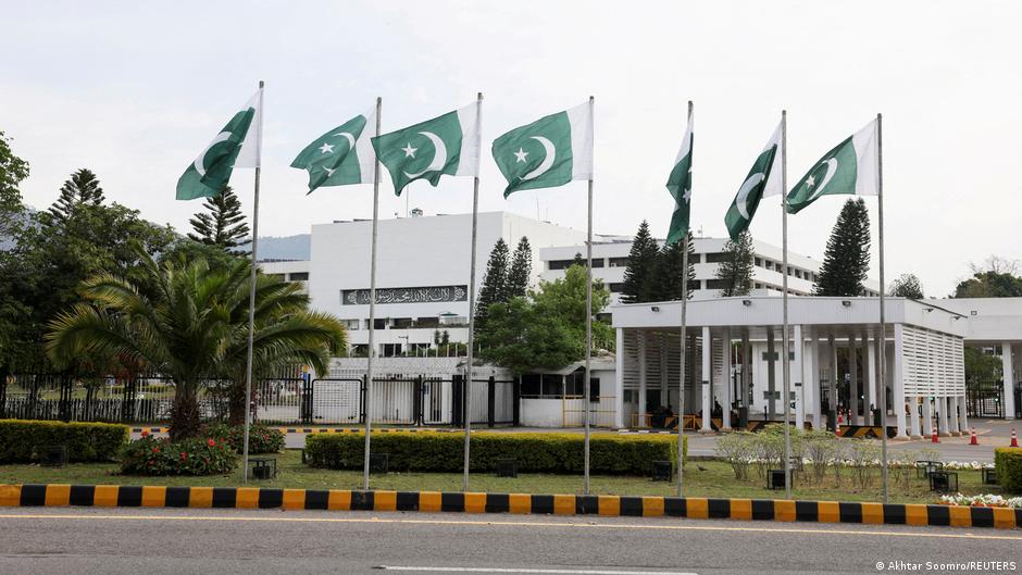 Pakistani flags flying outside a government building beside a guarded entrance and landscaped grounds