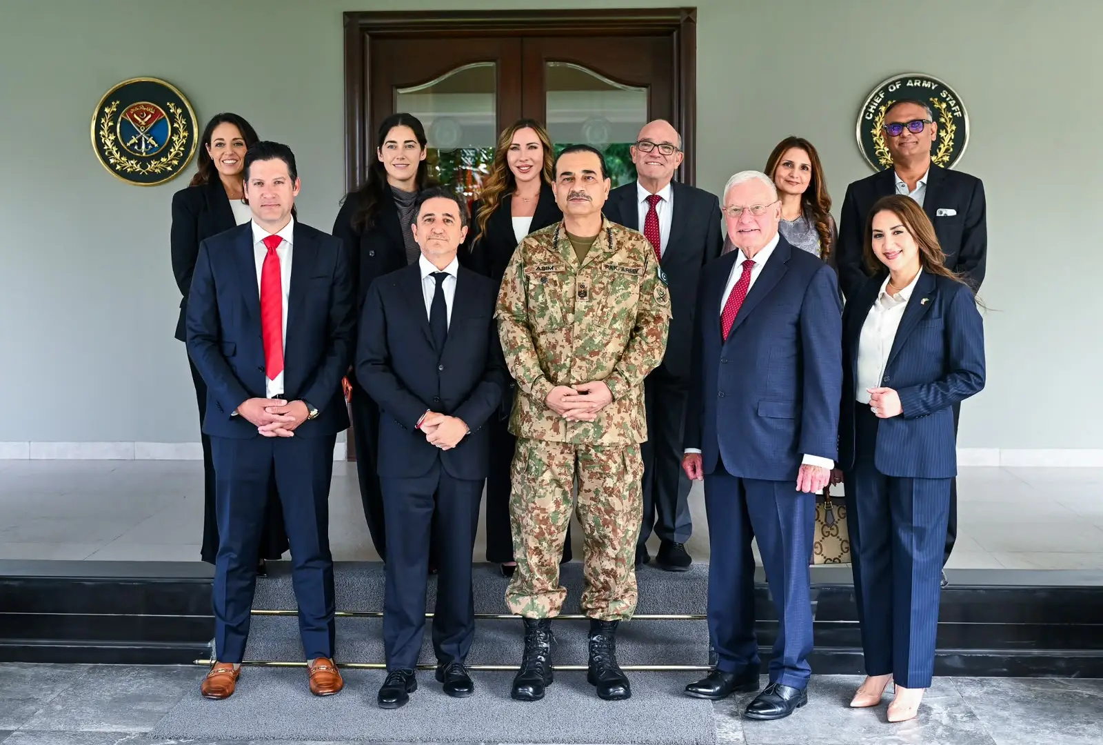 Officials and military personnel pose together on steps inside a building, with two military crests behind them.